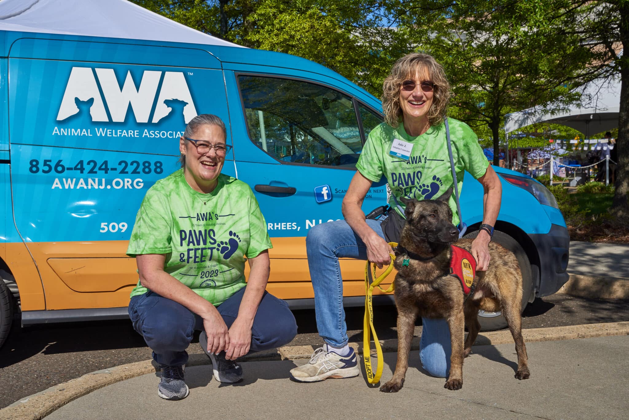 Two women in green shirts posing with shepherd dog in front of AWA Van.