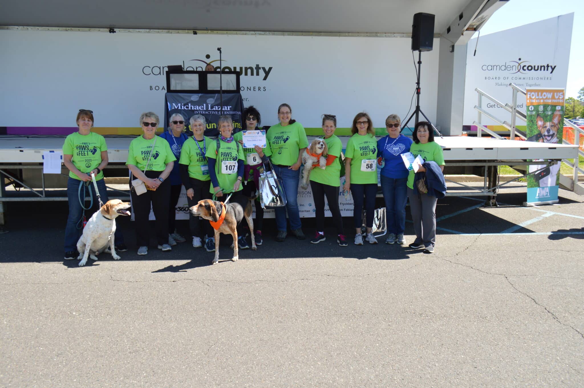 Group of women in green shirts with 2 dogs posing at Paws & Feet stage.