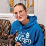 Caucasian woman with glasses and slicked back light hair holding a brown chihuahua wearing a pineapple jacket