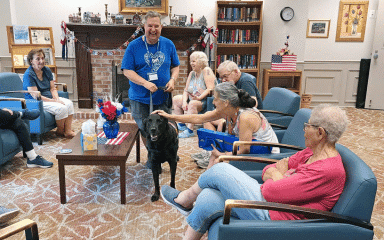Older, Caucasian man in a blue t-shirt with a black lab walking around an assisted living home for the elderly. The dog is receiving praise from one of the residents.