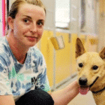 Caucasian woman with light brown hair, wearing a tie-dye tshirt, kneeling next to a tan dog with upright ears.