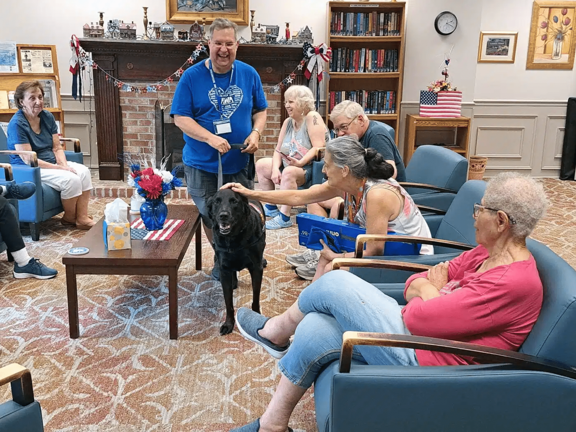 Man in blue shirt walking black labrador dog in senior home during pet therapy visit.