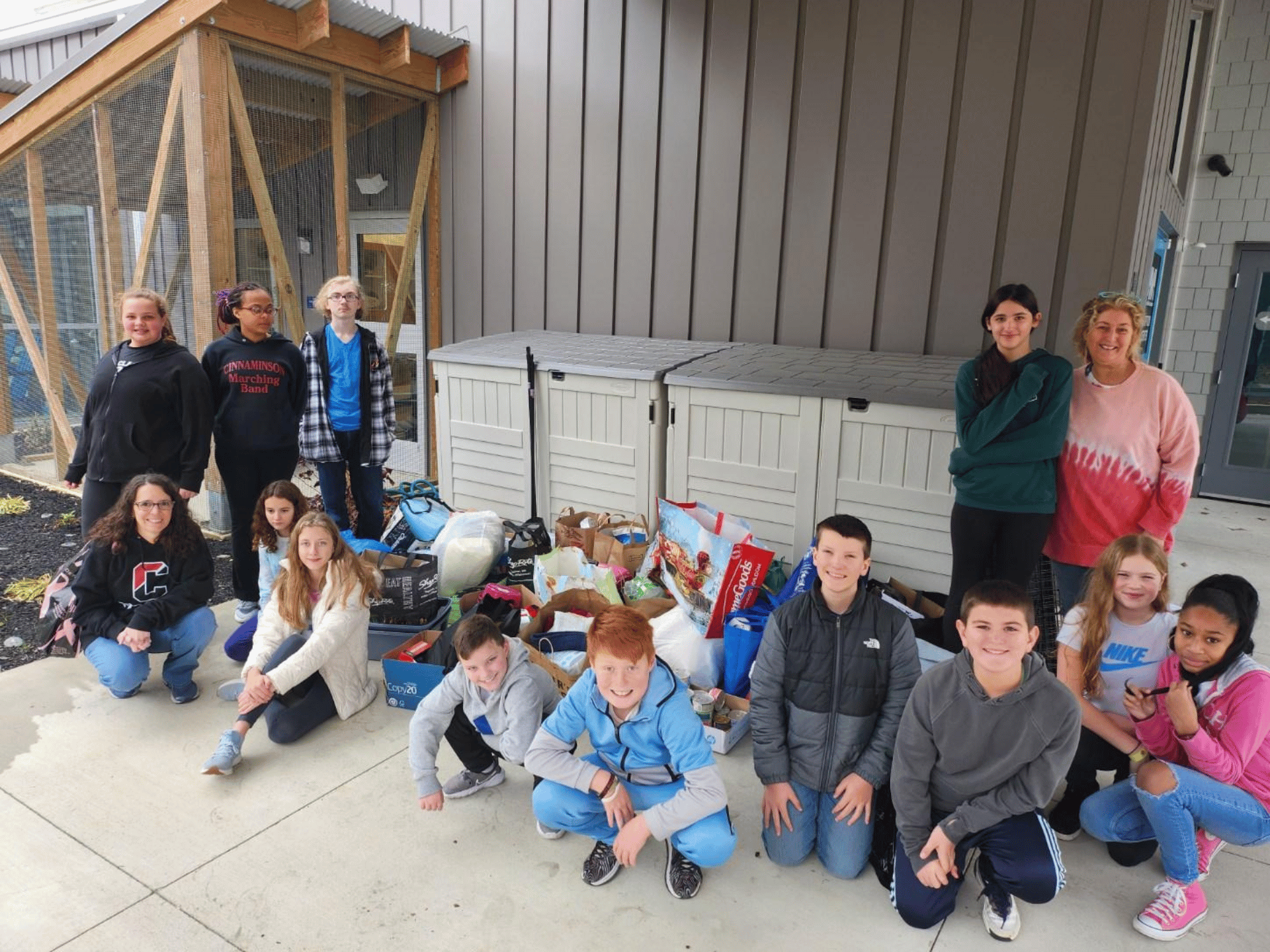 A group of school-aged kids sitting and standing in front of gray bins by a building