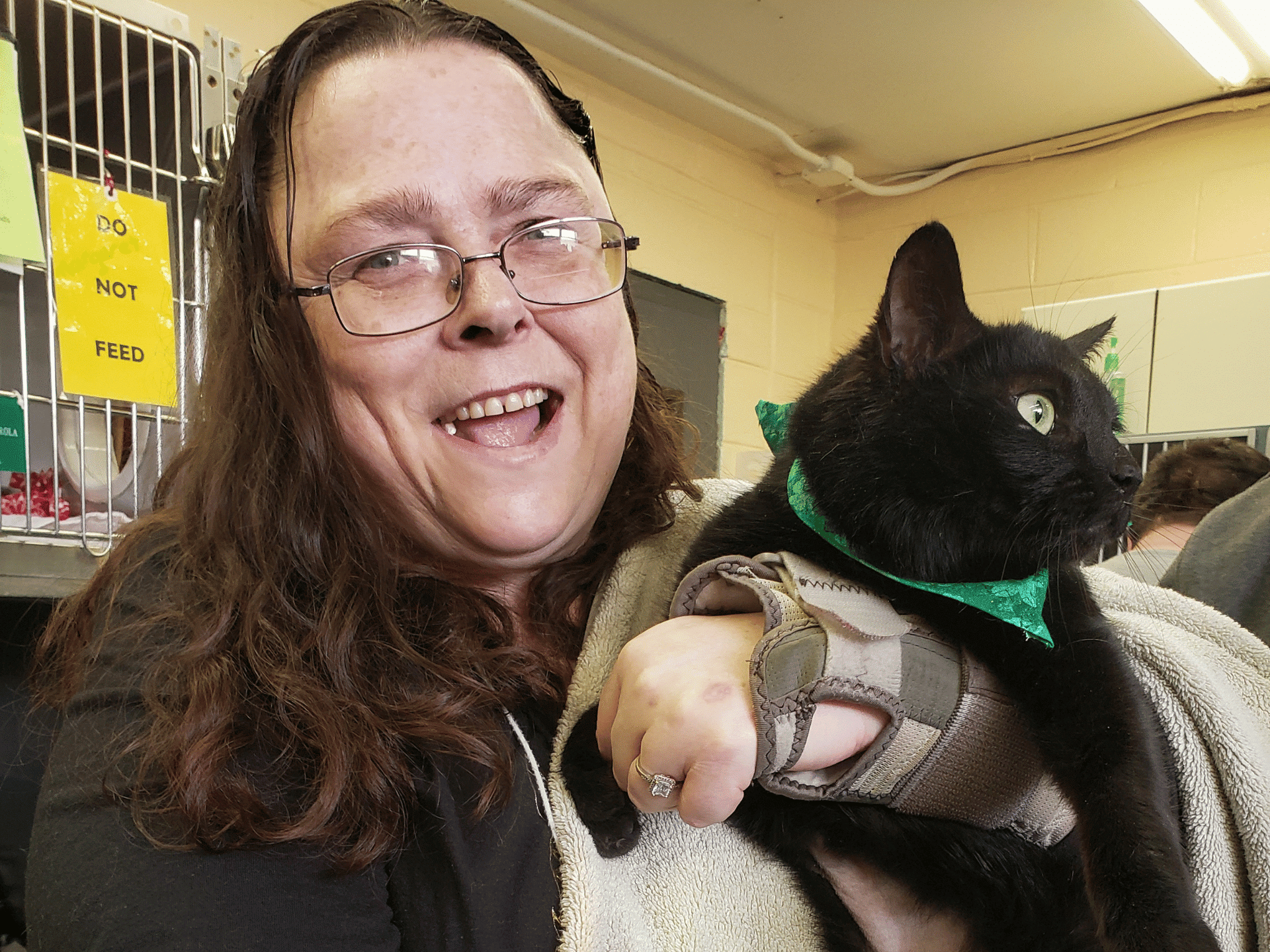 Caucasian woman with brown hair wearing glasses and a hand brace holding a black cat with green eyes